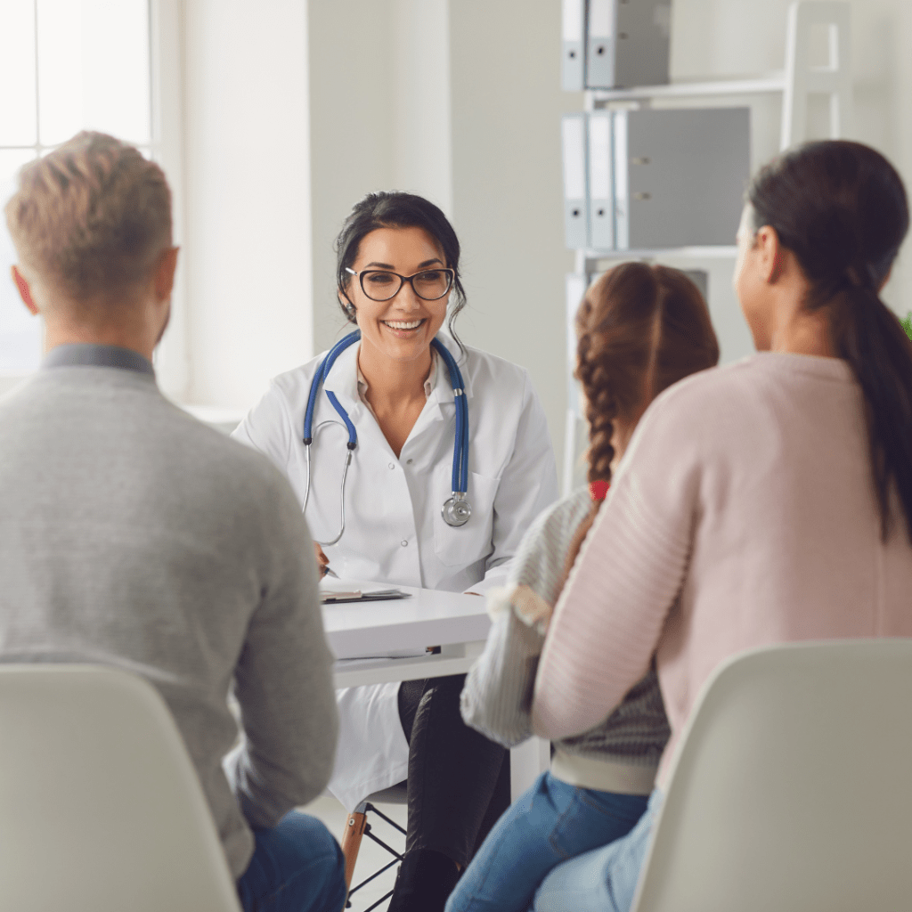 A female doctor wearing a stethoscope is talking to two adults and smiling. On the woman's lap is a child wearing her hair in plaits also looking at the doctor. 