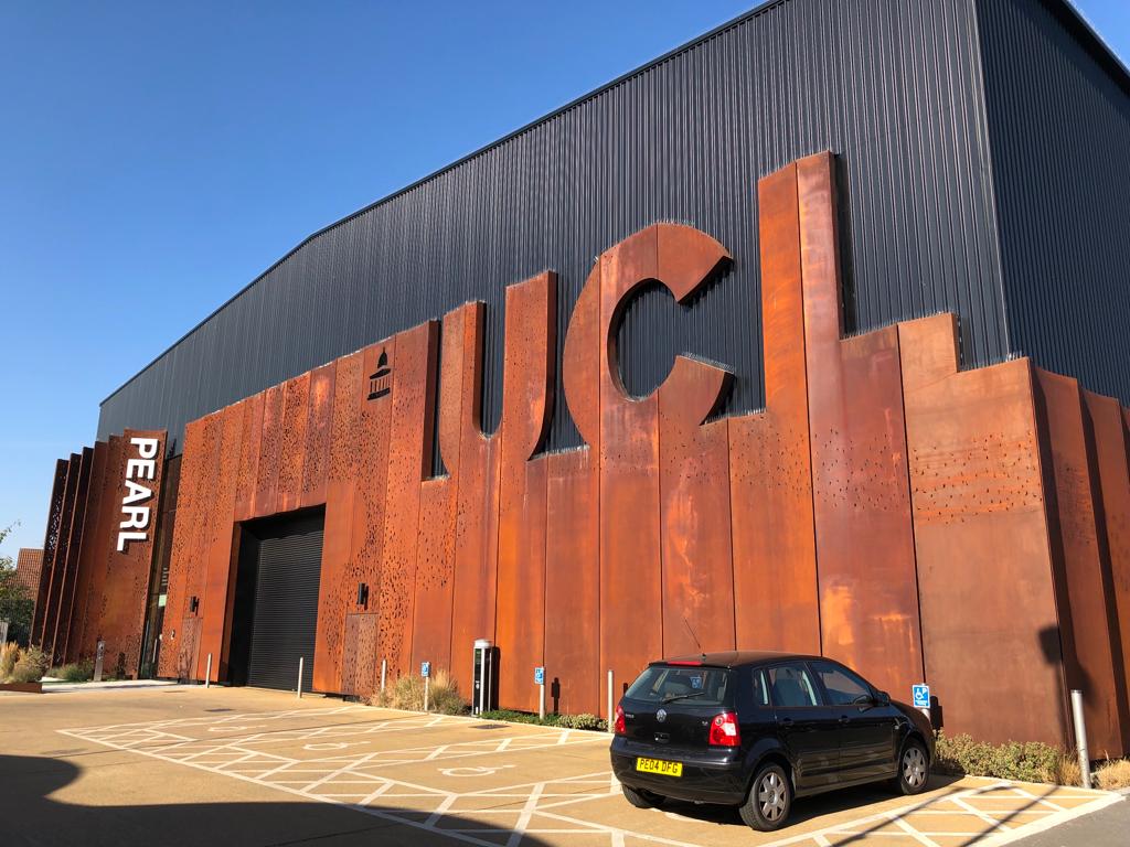 Image of a copper and black building with the word PEARL and UCL on it. There is a set of four disabled car parking spaces one of which has a car parked in it
